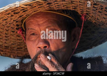 Xingping, Guangxi, China - 18 June 2026: View of an elderly man wearing a traditional woven straw hat while smoking a cigarette in Xingping Ancient To Stock Photo