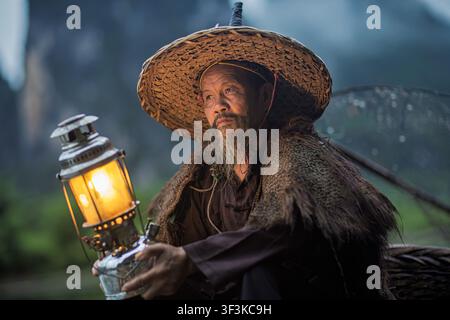 Xingping, China - 18 June 2026: View of an elderly fisherman wearing a traditional straw hat and holding a glowing lantern in Xingping Ancient Town, G Stock Photo