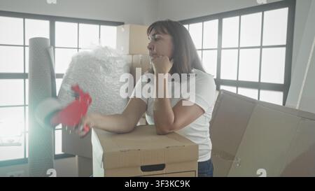 Woman holding tape dispenser, resting chin on fist in building with stacked boxes and bubblewrap; tired patience. Stock Photo