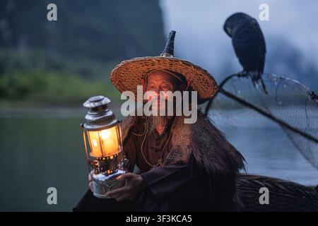 Xingping, Guangxi, China - 18 June 2026: View of an elderly fisherman in a traditional straw hat and cape holding a glowing kerosene lantern on a boat Stock Photo