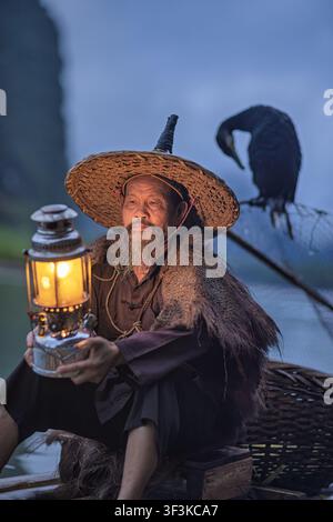 Xingping, China - 18 June 2026: View of a traditional cormorant fisherman holding a glowing lantern on a bamboo raft with his bird in Xingping Ancient Stock Photo