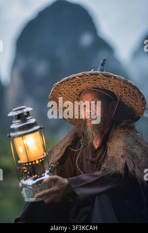 Xingping, Guangxi, China - 18 June 2026: View of an elderly fisherman wearing a traditional straw hat and holding a glowing lantern in Xingping Ancien Stock Photo