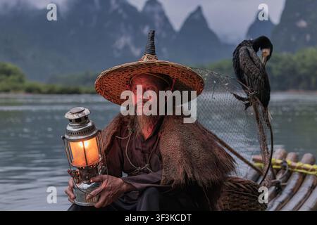 Xingping, Guangxi, China - 18 June 2026: View of a traditional cormorant fisherman holding a glowing lantern on a bamboo raft on the Li River with kar Stock Photo