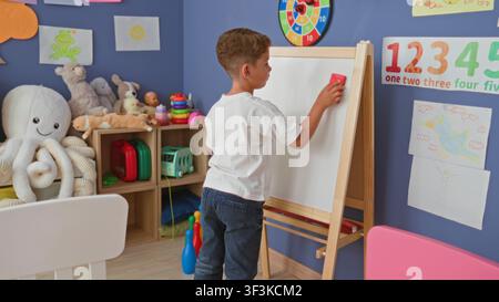 Boy standing erases easel whiteboard with red eraser in preschool classroom surrounded by stuffed toys and posters; concentration learning. Stock Photo