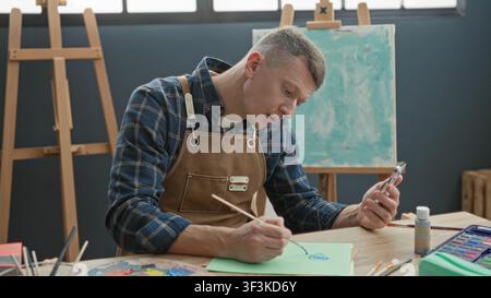 Man painter in apron holds smartphone in one hand and paints with brush on paper at an easel in studio with palette and water jar visible; concentrati Stock Photo