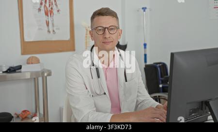 Man doctor typing on keyboard at desktop computer with stethoscope around neck, hands on keys, anatomical poster and brain visible on metal trolley in Stock Photo