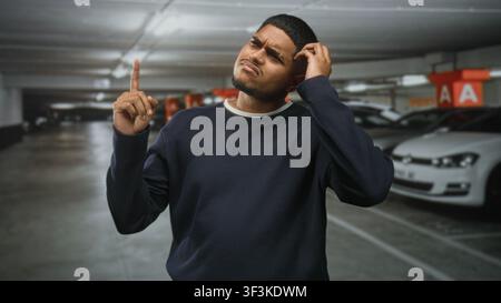 Hispanic man points finger up and scratches head in an underground parking building beside parked cars and concrete pillars marked a, looking puzzled; Stock Photo