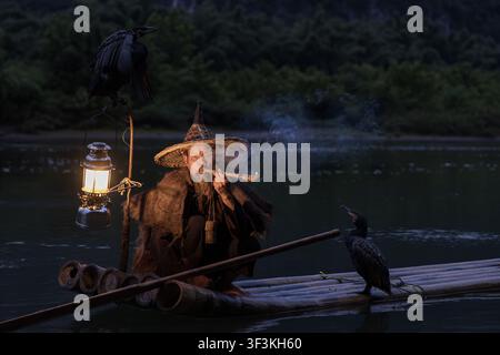 Guilin, Guangxi, China - 18 June 2026: View of an elderly fisherman smoking a pipe on a bamboo raft at night, with cormorants and a lantern on the Li Stock Photo