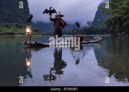Guilin, Guangxi, China - 18 June 2026: View of a traditional cormorant fisherman on a bamboo raft with birds, a lantern, and karst mountains in the ba Stock Photo