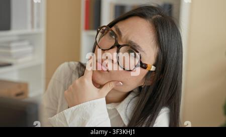 Woman wearing tortoiseshell glasses with hand on chin and pensive expression in office with shelves, binders and storage boxes; contemplation. Stock Photo