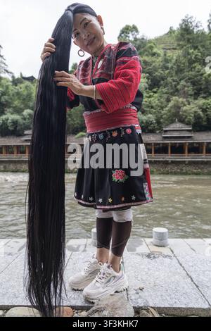 Guangxi, China - 18 June 2026: View of a Red Yao woman in traditional embroidered clothing holding her exceptionally long black hair, standing beside Stock Photo