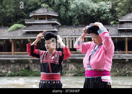 Guangxi, China - 18 June 2026: View of two Red Yao women in traditional clothing combing their long hair near a river with a traditional wooden buildi Stock Photo