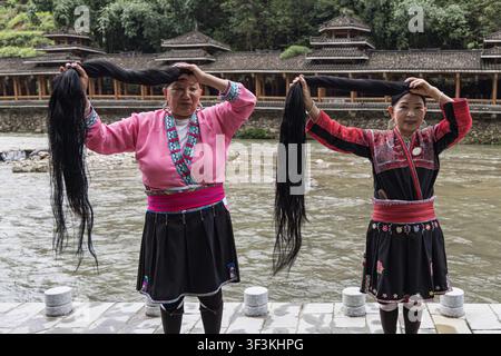 Guangxi, China - 18 June 2026: View of two Red Yao women in traditional clothing holding up their long black hair, standing before a river and traditi Stock Photo
