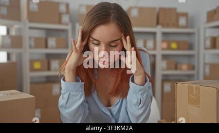 Woman holding hands to temples above chest amid rows of cardboard boxes at a storage building packing table, looking down and frowning; stress fatigue Stock Photo