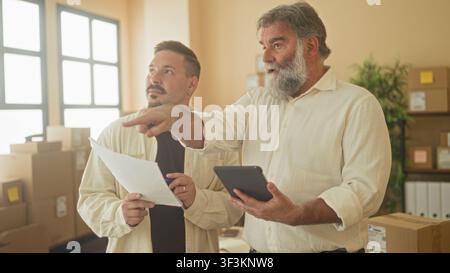 Two men examine parcel paperwork amid stacked boxes and shelving as a bearded man taps a tablet and younger man reads a sheet in a packing building; c Stock Photo