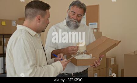 Two men inspecting a parcel, one writing on a clipboard while the other carefully holds an open box of packing filler amid stacked cartons in a storag Stock Photo