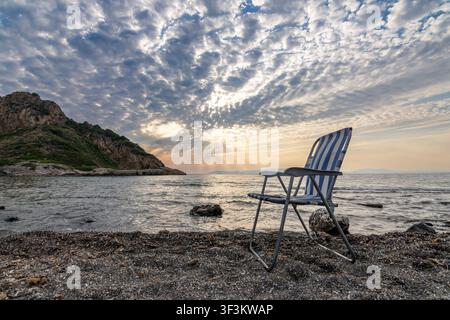 An empty striped lawn chair standing on a pebbled beach looking out to sea with sun rays bursting through clouds. Stock Photo