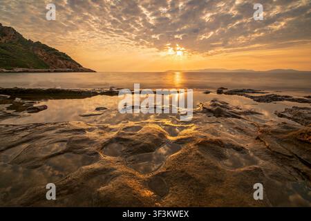 A vibrant sunset over the sea with golden sun beams filtering through clouds and reflecting off rock puddles along the shore. Stock Photo