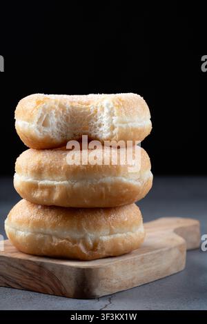 Close up of three glazed donuts stacked vertically on a wooden board, with a bite taken out of the top donut Stock Photo