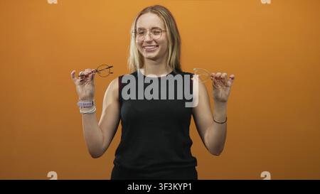 Woman holding two eyeglasses with hands raised, smiling while comparing frames against orange backdrop in studio; confident choice. Stock Photo
