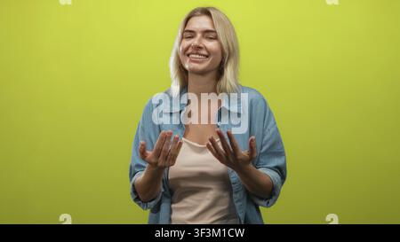Woman beckons with fingers and open hands, smiling in a denim shirt in a green studio; friendly invitation joy. Stock Photo