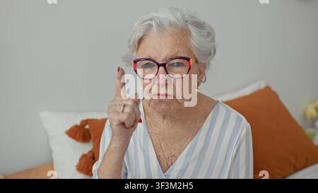 Elderly woman points finger while seated on bed in a bedroom building, wearing colorful glasses and pearl earring; disapproval. Stock Photo