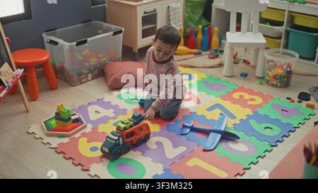 Boy seated on a colorful number mat holding a red toy truck with both hands, playing with cars and foam blocks in a playroom; quiet concentration. Stock Photo
