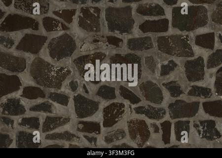 A rustic dark grey stone wall texture, showing irregular stones with visible mortar gaps. Stock Photo