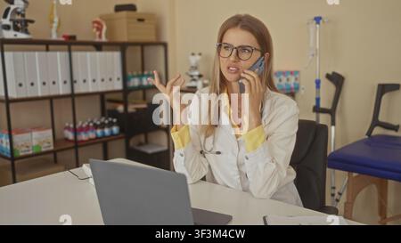 Woman doctor wearing white coat and stethoscope talking on phone, gesturing with hand over laptop in a medical building; telemedicine frustration. Stock Photo