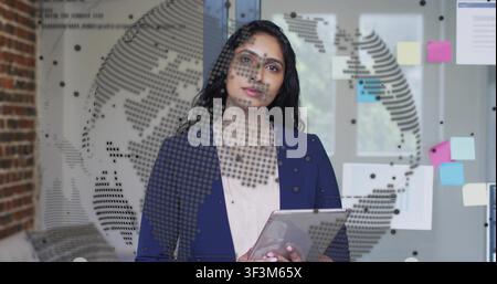Woman tapping tablet in office activating dotted-globe overlay and analyzing business data Stock Photo