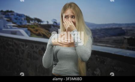Young blonde woman pinches nose with hand and covers mouth with other hand on building terrace by the sea, wearing gray long sleeve top; disgust odor. Stock Photo