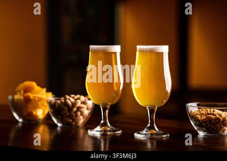 Two glasses with light beer and foam on wooden bar counter in interior of pub, pistachios, crackers and nuts in glass plates nearby Stock Photo