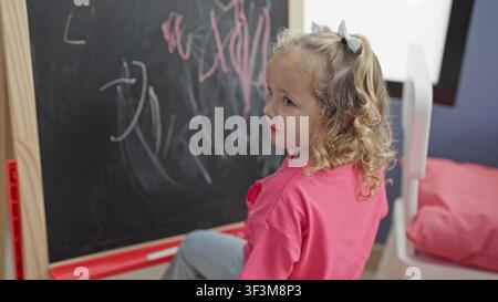 Girl drawing on chalkboard in a colorful playroom building, wearing pink shirt and turning toward mat and toys; curiosity learning play. Stock Photo