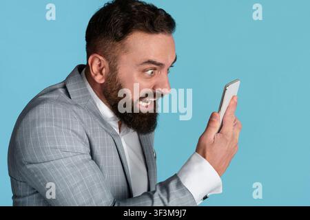 Angry young manager shouting at smartphone in fury on blue background Stock Photo