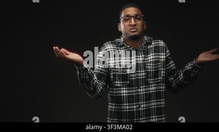 Young black man wearing glasses shrugs with palms up, open hands visible, plaid shirt, facing camera in studio under soft directional light; uncertain Stock Photo