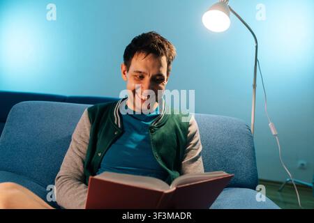 Happy man relaxing on blue couch under floor lamp, smiling while engrossed in novel, symbolizing leisure, learningfinding joy in literature indoors. Stock Photo