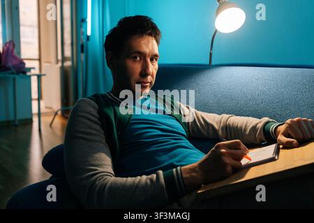 Creative man finding inspiration while sketching or writing in small notebook, sitting comfortably on blue sofa illuminated by lamp in cozy modern hom Stock Photo