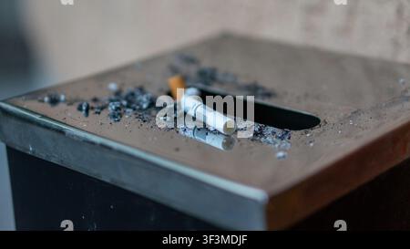 Ashes scatter near a cigarette on a metal ashtray. Smoke drifts softly from the spent cigarette. The ashtray's opening is dark and worn. This moment s Stock Photo