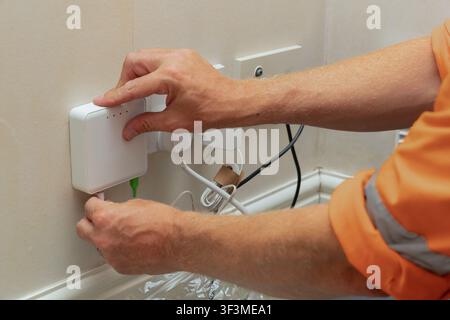 Telecoms engineer in hi-vis top plugging power cable into wall-mounted optical network terminal with SC APC fibre cable already connected, during resi Stock Photo