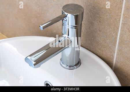 A close-up view of a sleek, contemporary chrome faucet with a single lever, installed on a pristine white ceramic sink with tiled background Stock Photo