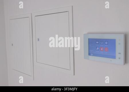 A sleek, white intercom panel with a blue touchscreen interface is installed on a clean, minimalist wall next to access panels Stock Photo
