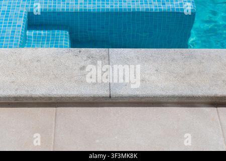 A detailed perspective of the poolside, showcasing the transition from textured stone coping to submerged mosaic tiles and clear blue water Stock Photo