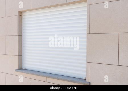A contemporary building exterior features a window covered by a closed white roller shutter, set against light beige stone cladding Stock Photo
