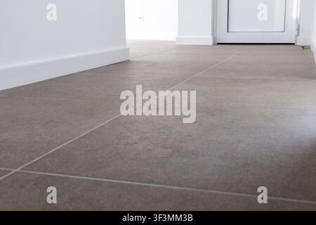 A contemporary hallway featuring large format grey stone tiles laid on the floor with clean white baseboards and a doorway in the background Stock Photo
