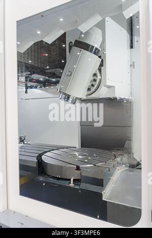 A close-up shot through protective glass of a modern 5-axis CNC milling machine chamber. heavy-duty rotary table, precise tilt-head spindle, and metal Stock Photo