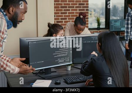 Mixed programmer team works on a group software project across multiple screens, with a coder pointing at code while discussing development tasks in an IT office. Stock Photo