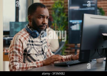 Concentrated portrait with african american programmer wearing wireless headphones working at computer screen while typing at a computer, showing a focused developer workflow with code, headphones. Stock Photo