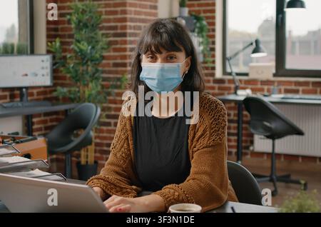 Masked businesswoman using laptop in startup in startup office while working through documents, digital reports and company planning tasks. Stock Photo