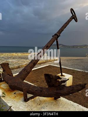 Large rusted ship anchor displayed at Artemida Harbor in Attica, Greece, with the sea and coastal settlement in the background. Stock Photo