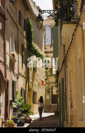 A woman walks her dog down a narrow, sunlit historic alley lined with traditional Provencal buildings in Arles, France. Stock Photo